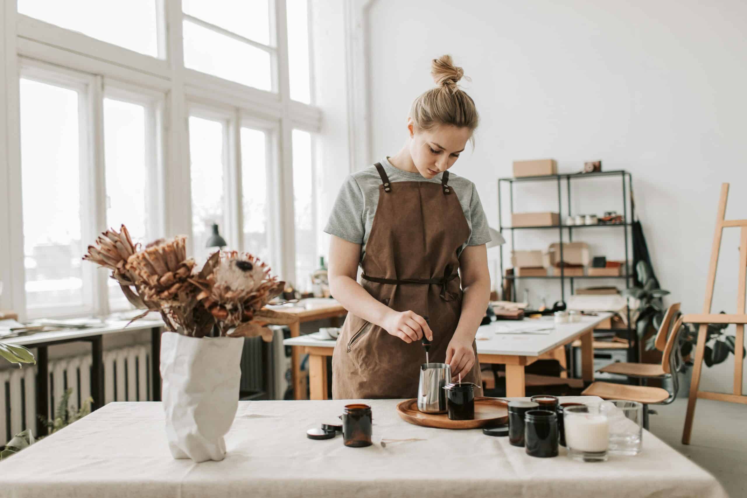 Person in a brown apron decorating at a well-lit studio table with dried flowers in the foreground, concentrating on a task.